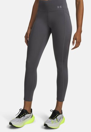 Persoon met grijze sportlegging met hoge taille en witte hardloopschoenen met neongele zolen, staand tegen een effen achtergrond.