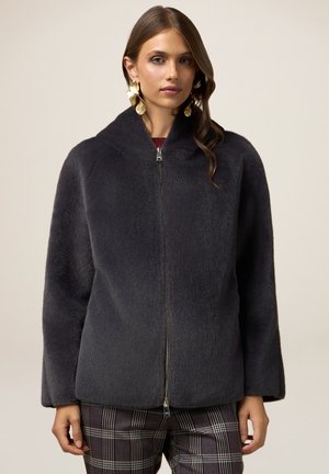 Giacca grigia carbone con zip frontale, realizzata in un morbido tessuto testurizzato, con colletto rotondo e spalle scese; abbinata a pantaloni a quadri.