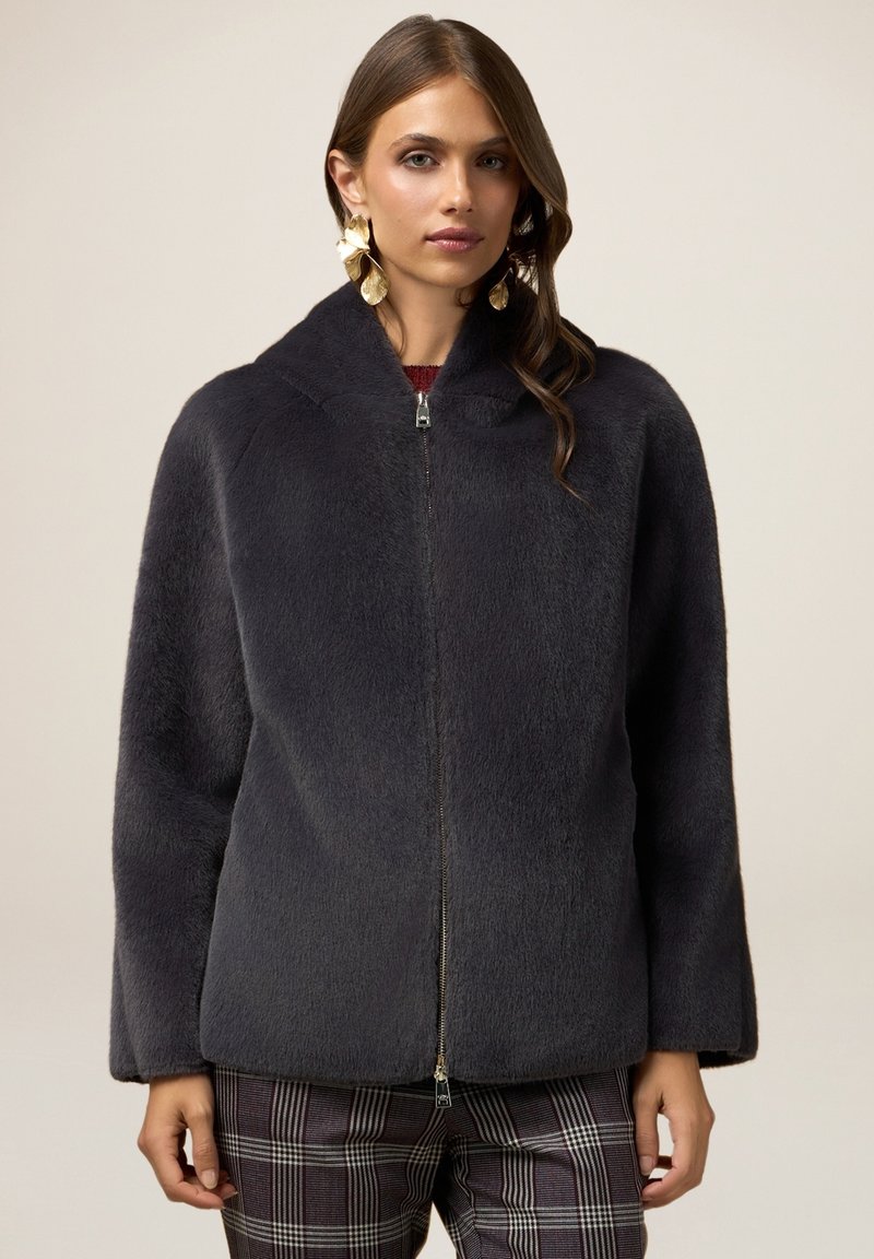 Chaqueta gris carbón con cremallera frontal, que presenta un tejido suave y texturizado, cuello redondo y hombros caídos; combinada con pantalones de cuadros.