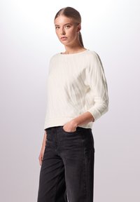 Maglione bianco testurizzato con colletto rotondo e maniche a tre quarti, abbinato a jeans neri a vita alta. Design semplice e moderno.