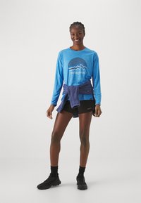 Blå langærmet skjorte med et cirkulært grafisk design og teksten "Patagonia", parret med sorte shorts og en blå sweater bundet om livet.
