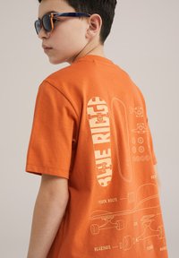 Pomarańczowy bawełniany t-shirt z nadrukiem deskorolki na plecach, z białymi grafikami przedstawiającymi elementy deskorolki oraz tekst.