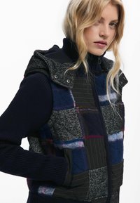 Gilet imbottito con cappuccio, realizzato in un mix di tessuti blu scuro e a scacchi, pannelli testurizzati e tasche laterali. Accenti di cucitura robusta.