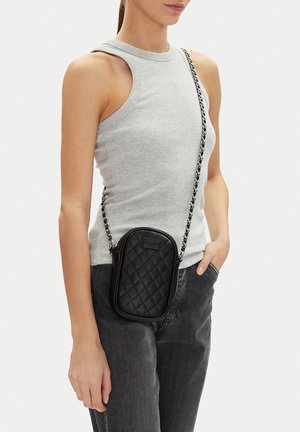 Žena v šedém tílku a tmavých džínách, nesoucí malou černou prošívanou kabelku přes rameno s řetízkovým popruhem.