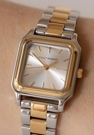 Montre bracelet carrée en argent et en or avec un cadran réfléchissant, des index horaires dorés et un bracelet en métal bi-ton avec des maillons polis.