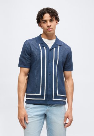 Jeune homme aux cheveux bouclés portant une chemise bleu marine à manches courtes avec des rayures blanches verticales et un jean bleu clair.