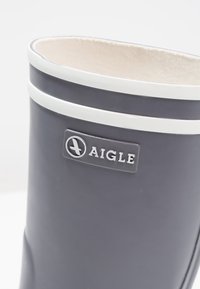 Botte en caoutchouc grise avec des rayures blanches et un patch logo portant le mot "Aigle". L'intérieur est doublé d'un matériau clair.