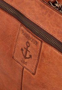 Braune Leder Tasche mit einem Reißverschlussverschluss und einem Markenpatch mit einem Ankerdesign. Das Leder hat eine strukturierten Oberfläche.