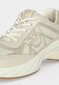 Gros plan d'une basket beige et blanche avec des lacets blancs, un tissu texturé et un logo oiseau en relief sur le côté et la languette.