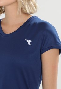 Navy blue short-sleeve athletic top made from smooth fabric, featuring a white logo on the left chest and a wide crew neckline.