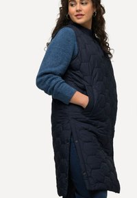 Vatterad marinblå väst med hexagonmönster, sidofickor och tryckknappar. Bärs över en blå texturerad långärmad tröja och jeans.