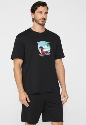 Mann trägt ein schwarzes T-Shirt mit einer Grafik eines Dackels auf einem Skateboard und dem Schriftzug „Enjoy the Ride“, kombiniert mit schwarzen Shorts.