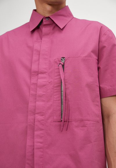 Camicia a maniche corte in cotone magenta con colletto a bottoni e taschino con zip, dotato di accessori in argento. Texture liscia.