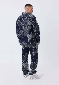 Hombre de pie de perfil, vistiendo una chaqueta de cuadros pixelados en azul oscuro y gris, junto con pantalones a juego y zapatillas blancas.