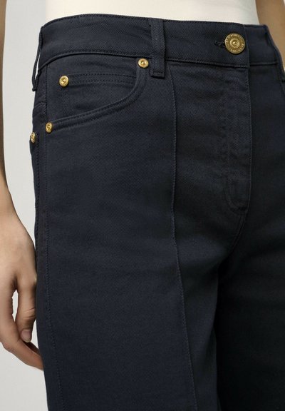I jeans in denim blu scuro presentano una vestibilità aderente, rivetti in tonalità oro e una chiusura con bottone. Il tessuto ha una texture liscia con una leggera elasticità.