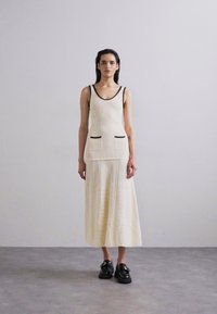 Top sin mangas de raya en crema con detalles en negro y bolsillos, combinado con una falda larga a juego, llevado con zapatos negros.