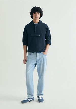 Joven de pie con una mano en el bolsillo, vistiendo una sudadera con capucha azul marino, vaqueros azul claro y zapatillas azules sobre un fondo liso.