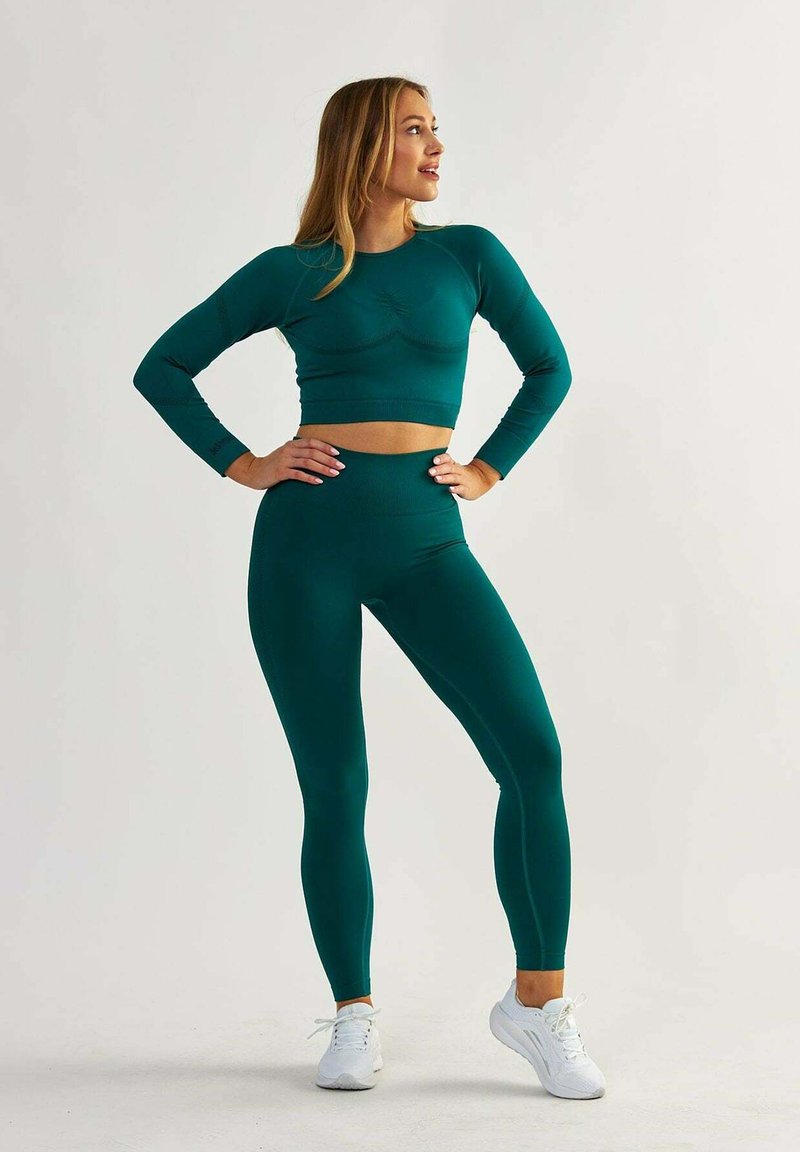 Zestaw do ćwiczeń w kolorze niebiesko-zielonym składający się z krótkej bluzki z długim rękawem i legginsów o wysokim stanie, wykonany z gładkiego, elastycznego materiału, w połączeniu z białymi butami sportowymi.