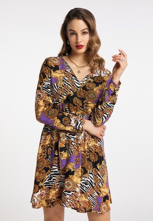 Vrouw in een kleurrijke jurk met patronen en gouden, zwarte, paarse en witte details, poseert met één hand omhoog en de andere gekruist bij de taille.
