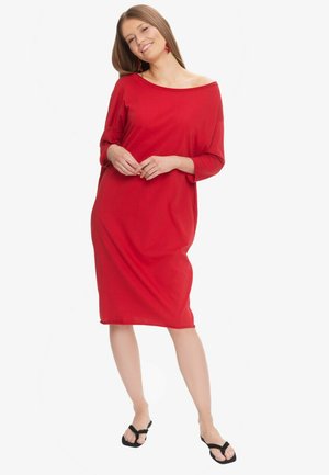 Uśmiechnięta kobieta stojąca, ubrana w luźną czerwoną sukienkę z rękawami za łokieć i czarne klapki na białym tle.