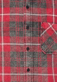 Camicia a quadri rossa e grigia con chiusura frontale a bottoni neri e taschino sul petto. Il tessuto presenta un aspetto strutturato.