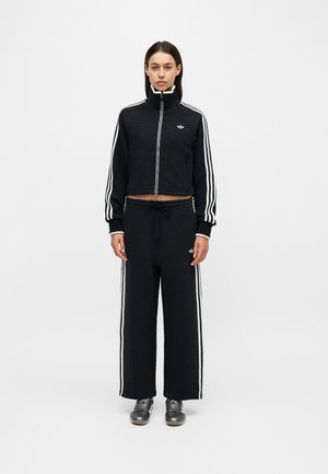 Vrouw staat naar voren gericht en draagt een zwart Adidas trainingspak met witte strepen, broek met wijde pijpen en grijze sneakers tegen een effen achtergrond.