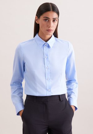 Kobieta o długich ciemnych włosach, ubrana w jasnoniebieską koszulę na guziki i czarne spodnie, stoi z rękami w kieszeniach na jednolitym tle.