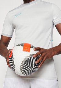 Šedé sportovní tričko Nike s krátkým rukávem, se smaragdově modrým logem. Osoba drží vzorovaný fotbal s černými a oranžovými akcenty.