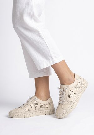 Jambes portant un pantalon blanc court et des baskets beiges perforées avec des semelles texturées sur un fond blanc uni.