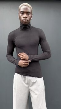Mlad moški s kratkimi blond lasmi, oblečen v črn turtleneck in svetlo sivo hlače, stoji ob enostavni sivi steni in prilagaja rokav.