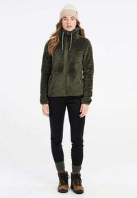 Groene fleece jack met hoge kraag en ritssluiting, gecombineerd met zwarte slim-fit broeken en bruine laarzen. Model draagt een beige gebreide beanie.