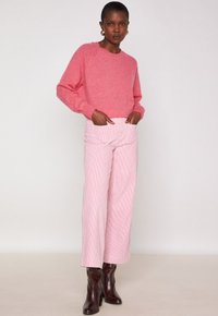 Donna in piedi con le mani in tasca, indossa un maglione rosa, pantaloni a palazzo a righe rosa chiaro a vita alta e stivaletti con tacco marroni scuri.