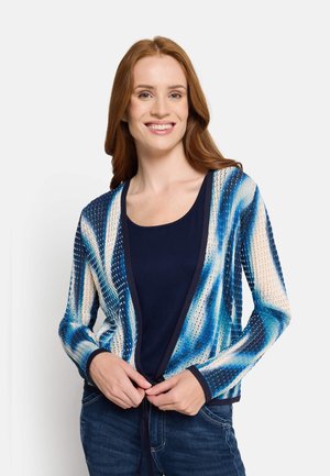 Young woman smiling, wearing a blue patterned open-knit cardigan over a navy shirt with hands adjusting the cardigan at waist level.