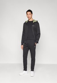 Μαύρη αθλητική φόρμα με πράσινο κουκούλα, με εφαρμοστή σχεδίαση και κεντημένο λογότυπο. Συνδυασμένη με λευκά αθλητικά παπούτσια. Μαλακή υφή υφάσματος.