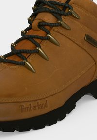 Καφέ δερμάτινο αναρριχητικό μποτάκι Timberland με μαύρα και γκρι κορδόνια περασμένα μέσα από μπρούτζινα κρικάκια και χοντρή μαύρη σόλα.