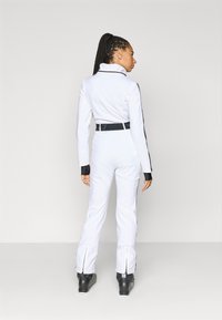 Λευκή ολόσωμη φόρμα με μαύρες λεπτομέρειες, ψηλό γιακά, εφαρμοστή σιλουέτα και φερμουάρ στα μανίκια. Συνδυάζεται με μαύρες μπότες σκι για εξωτερική χρήση.