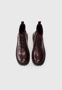 Botas de tobillo de cuero burdeos con cordones negros, suelas de goma contrastantes y lazos en la parte trasera; textura suave con detalles mínimos.