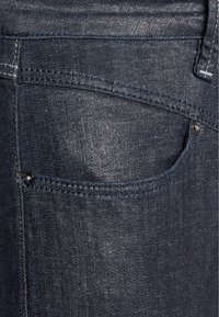 Ciemnoniebieskie dżinsy z fakturowanej tkaniny, z tylną kieszenią i dekoracyjnym przeszyciem. Zawierają metalowy nit akcentujący.