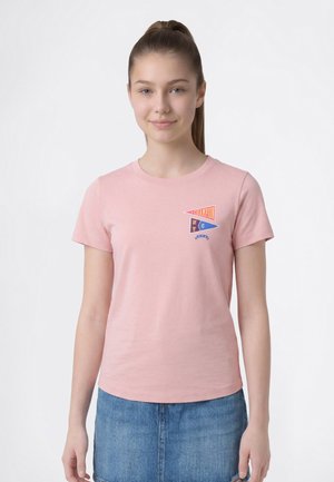 Mujer joven con cabello castaño recogido en una cola de caballo, lleva una camiseta rosa con pequeños diseños de banderines de colores y una falda vaquera azul.