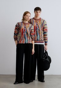 Pulls en tricot colorés avec des motifs ondulés, associés à de larges pantalons noirs. Un modèle tient un sac circulaire noir, tous deux portent des accessoires minimalistes.