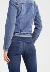 Giacca di jeans azzurro chiaro con design aderente, caratterizzata da doppie cuciture e chiusure con bottoni. Abbinata a jeans blu scuro, con tasche posteriori distintive.