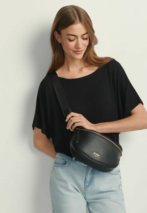 Kobieta ubrana w czarną bluzkę i jasnoniebieskie jeansy trzyma czarną skórzaną torebkę na ramię z złotym suwakiem i logo.