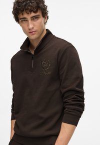 Hnedá zipsová mikina s textúrovanou tkaninou, s vysokým golierom a vyšitým logom na hrudi. Dlhé rukávy s rebrovanými manžetami.