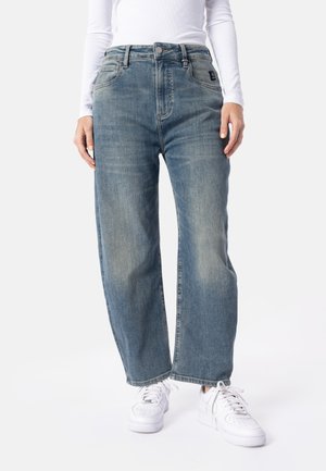 Person iført falmede blå jeans med lige ben, hvid langærmet top og hvide snøresneakers, stående mod en ensfarvet baggrund.