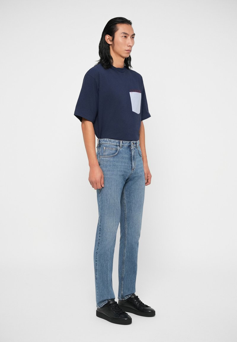 Mann mit langen schwarzen Haaren, der ein navyfarbenes T-Shirt mit einer hellblauen Tasche, hellblaue Jeans und schwarze Turnschuhe trägt, vor einem weißen Hintergrund steht.