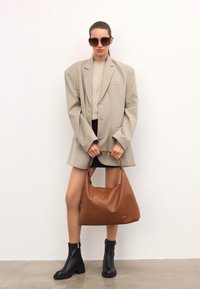 Mujer que lleva una chaqueta beige oversize, una falda corta negra, botas negras hasta el tobillo y gafas de sol, sosteniendo un gran bolso de cuero marrón.