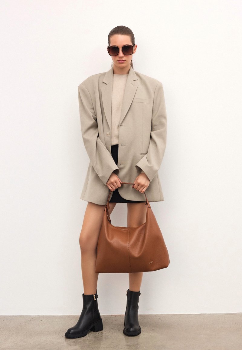 Mujer que lleva una chaqueta beige oversize, una falda corta negra, botas negras hasta el tobillo y gafas de sol, sosteniendo un gran bolso de cuero marrón.