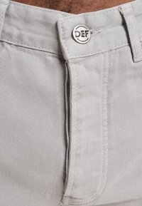 Light gray denim pants feature a metal button with "DEF" engraving, with visible stitching and a smooth texture. Close-up of the waistband area.