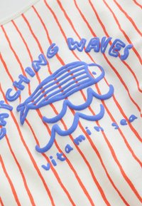 Poisson bleu brodé et vagues avec le texte "Catching waves" et "vitamin sea" sur un tissu blanc à rayures verticales orange.