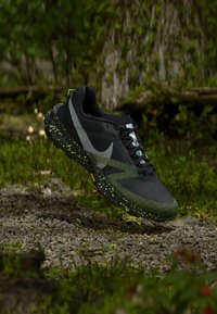 Schwarz-grüner Trailrunning-Schuh mit reflektierendem Swoosh, in der Luft über einem kieseligen und moosbedeckten Waldboden.